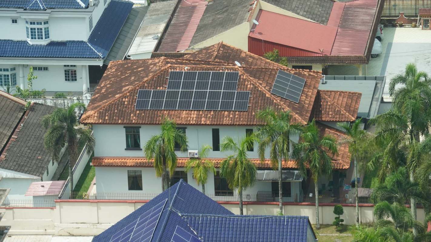 A residential building with a tiled roof and solar panels, surrounded by palm trees and other rooftops.