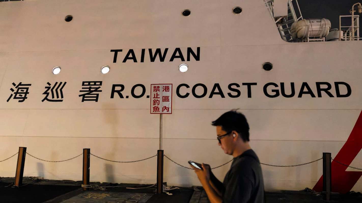 A man looks at his phone near a ship labeled "TAIWAN R.O.C. COAST GUARD" and a warning sign.