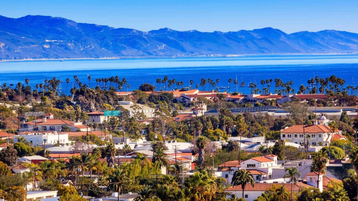 Santa Barbara, California, with red-tiled rooftops and palm trees, overlooking the ocean and mountains in the background.