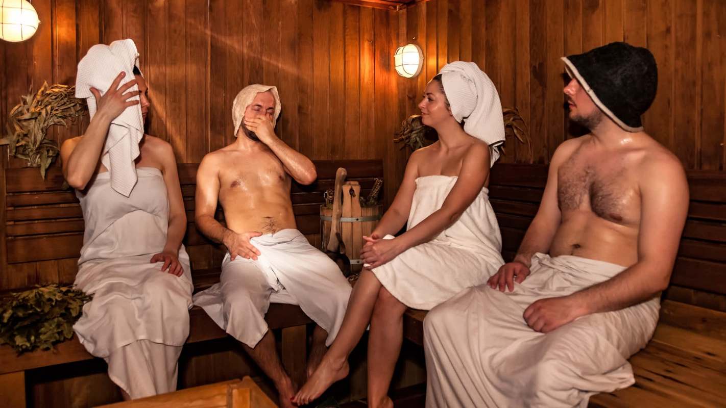 Four people relax in a sauna, wearing towels and sitting on wooden benches.