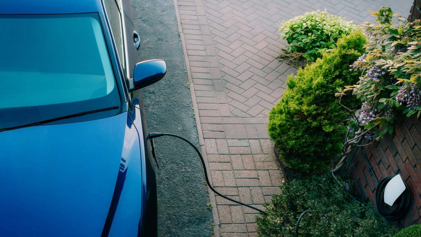 An electric vehicle is plugged in next to a paved walkway and plants.