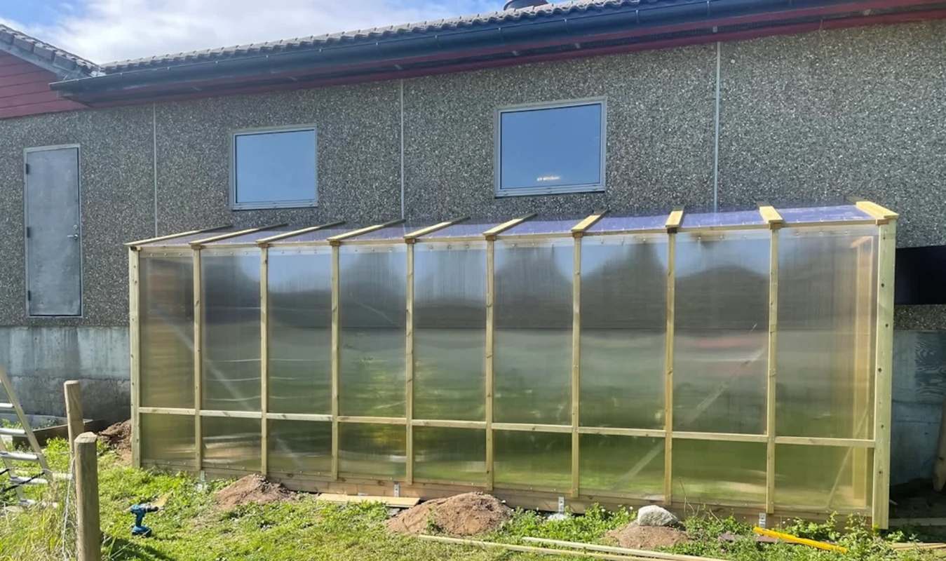 A greenhouse made of transparent panels and wooden framing beside a building.