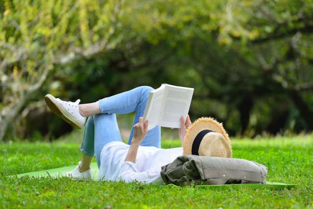 A person reading a book while lying on grass, wearing a straw hat and using a bag as a pillow.