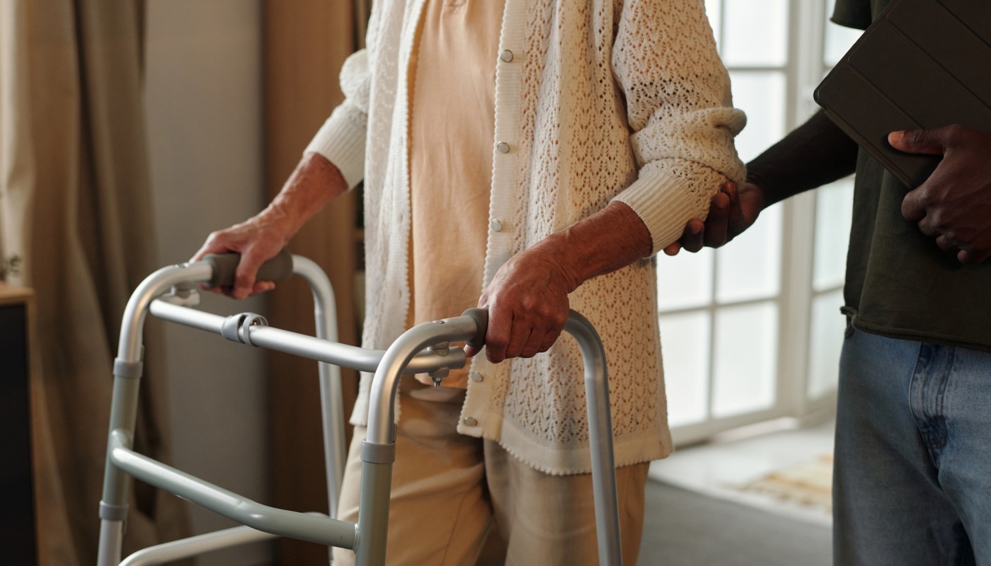 An older adult uses a walker while being assisted by another person.