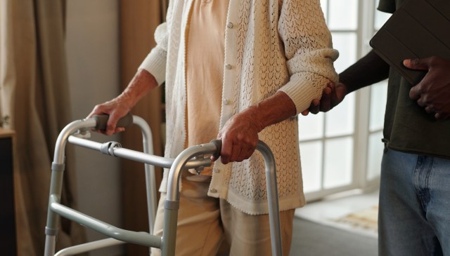An older adult uses a walker while being assisted by another person.