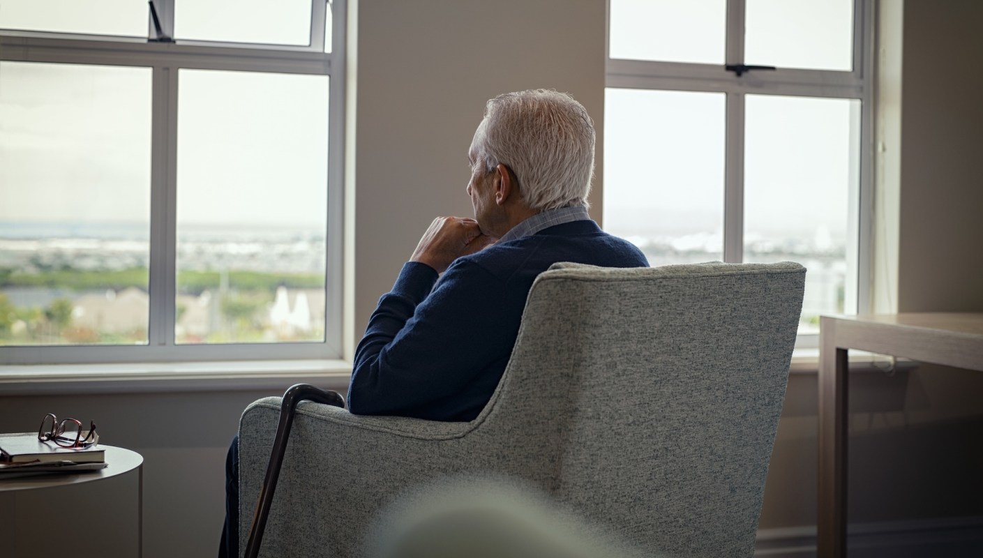 An older man seated in an armchair, looking out a window at a distant landscape.