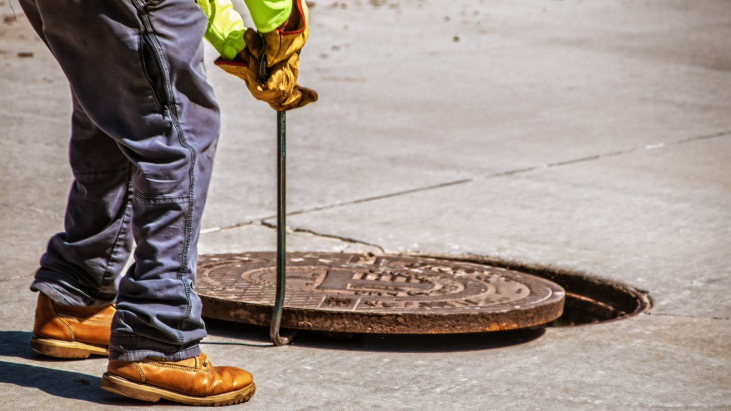 A person in work clothing uses a tool to lift a manhole cover on a pavement.