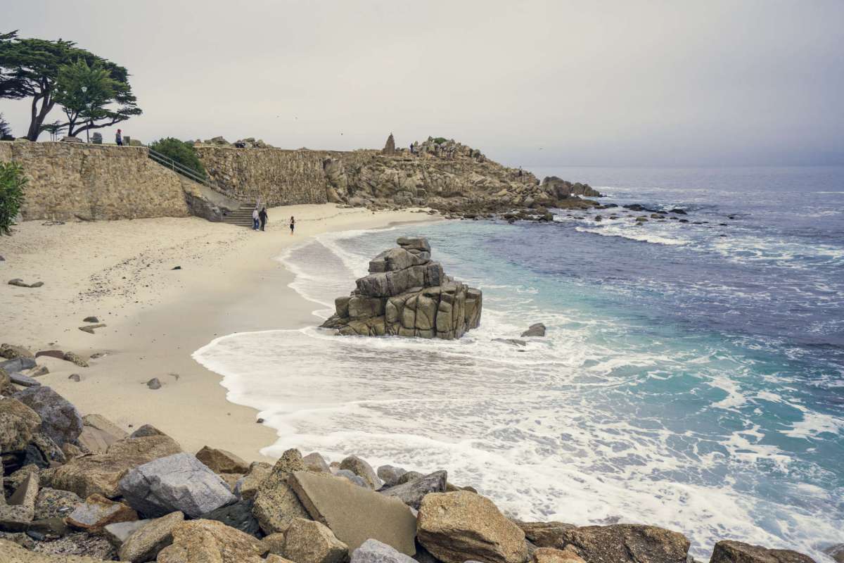 A rocky beach with waves, a stone wall, and people walking along the shore.