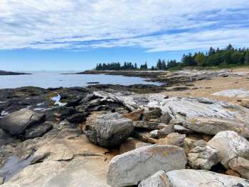A rocky shoreline with a calm sea and trees in the background under a partly cloudy sky.