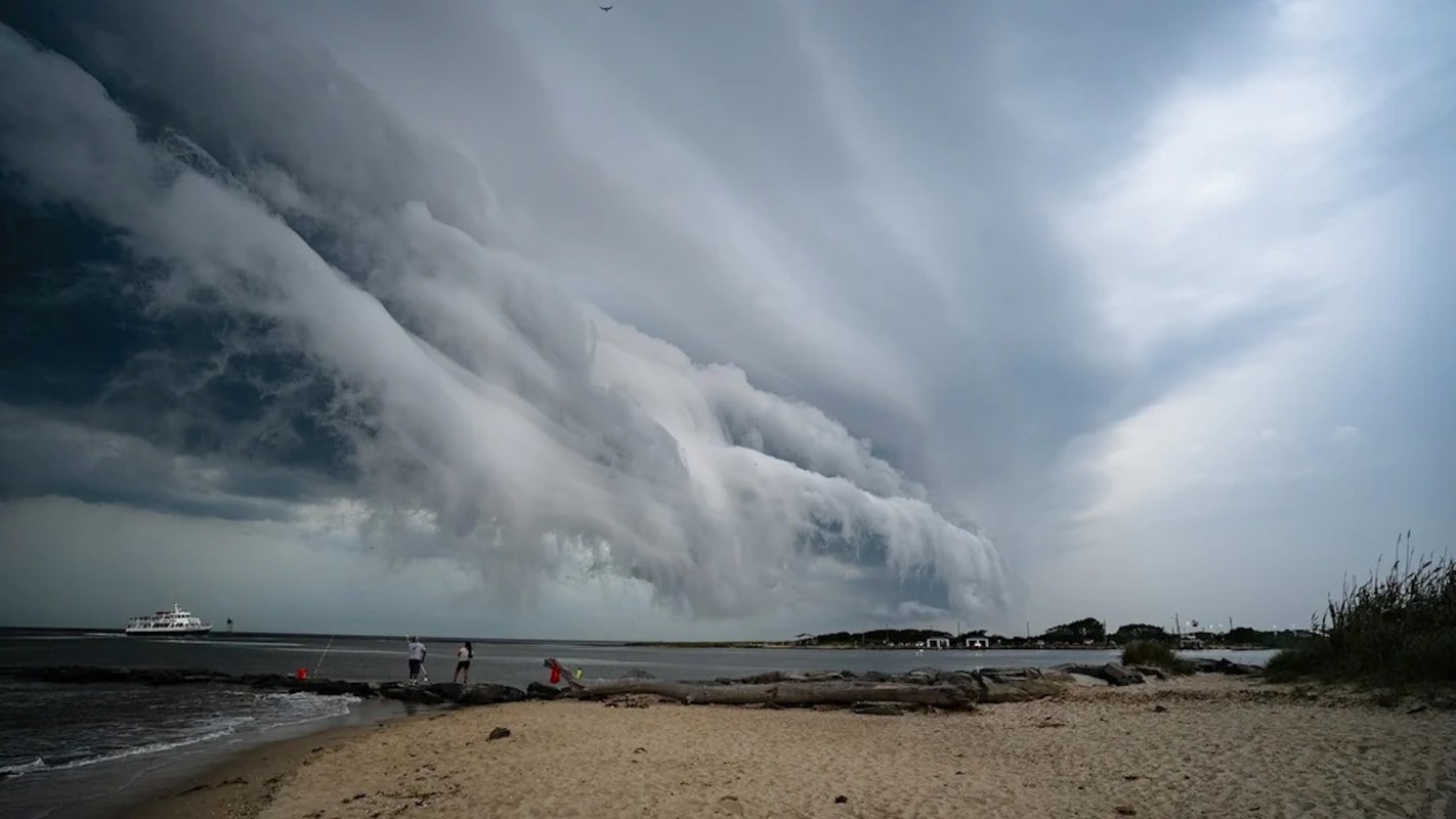 A beach scene with dark, rolling clouds that appear to form an overhanging shelf, with people fishing near the shore.