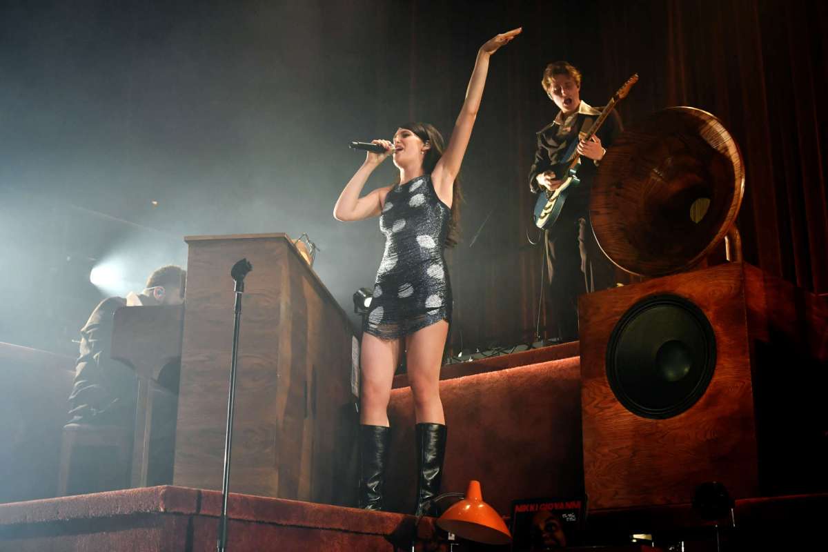 A singer performs on stage with musicians and vintage audio equipment in the background.