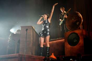 A singer performs on stage with musicians and vintage audio equipment in the background.