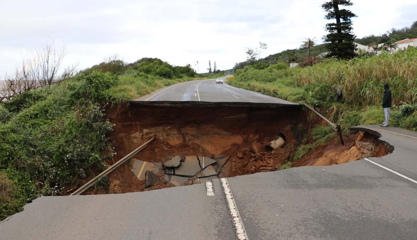A section of road is destroyed by a large sinkhole, with a person standing nearby observing the damage.