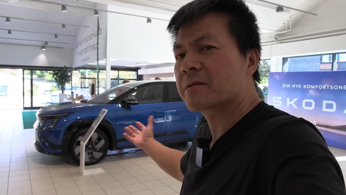 A man gestures towards a blue car displayed in a showroom with promotional signage.