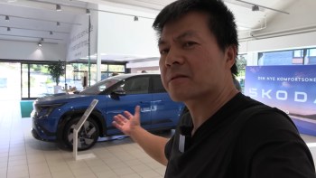 A man gestures towards a blue car displayed in a showroom with promotional signage.