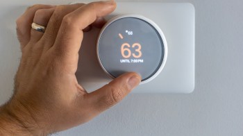 A hand adjusts a digital thermostat displaying a temperature of 63 degrees.