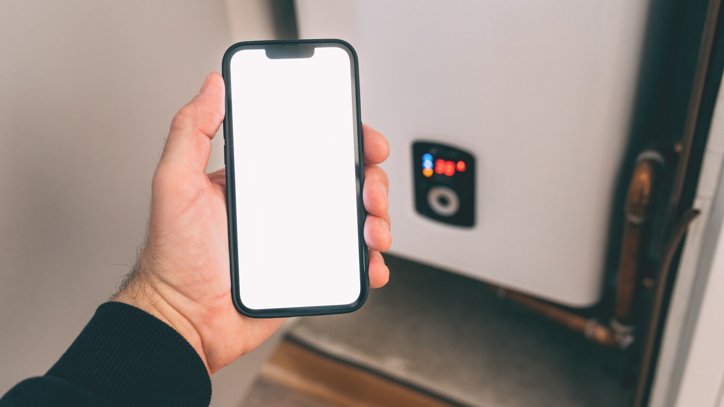 A hand holds a smartphone in front of a white appliance with a digital display.