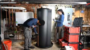 Two technicians work on a large black water heater in a basement filled with tools and piping.