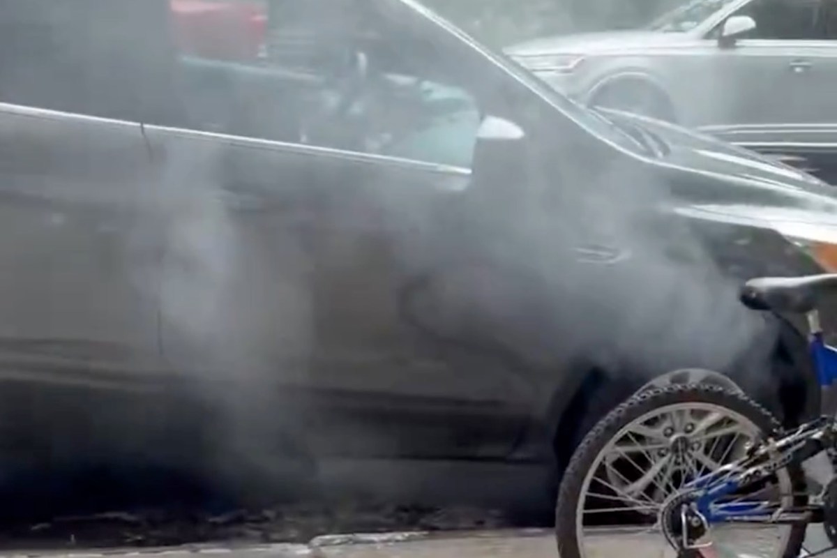 Smoke emanates from a parked black vehicle on a street, with a bicycle partially visible in the foreground.