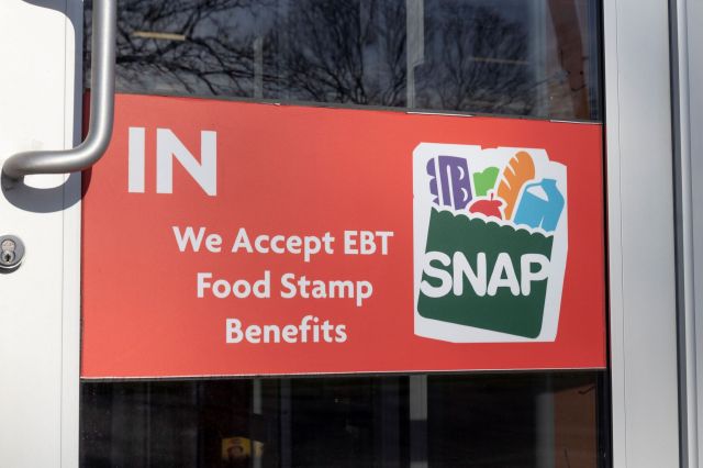 A sign on a door that reads "We Accept EBT Food Stamp Benefits" with a graphic of food items.
