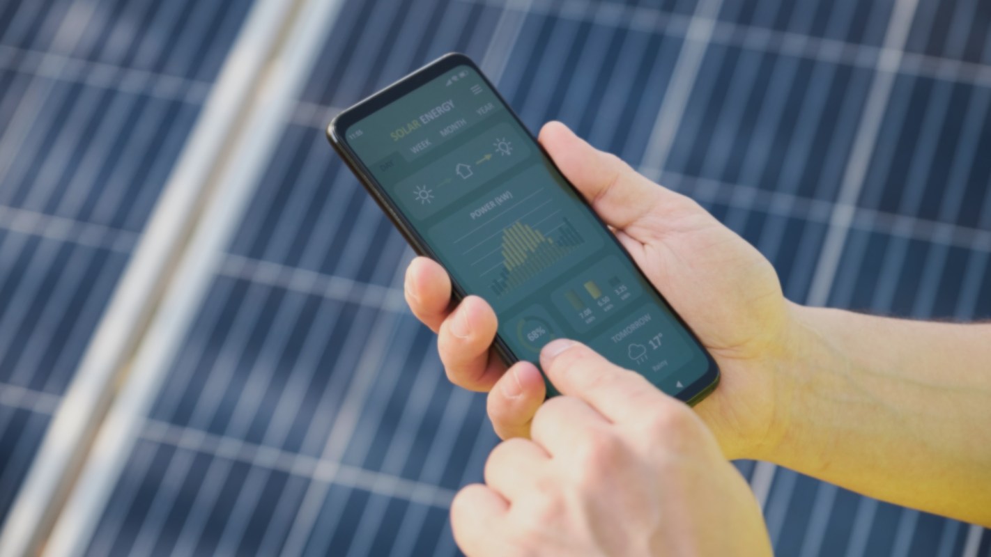 A person using a smartphone to monitor solar energy data with solar panels in the background.