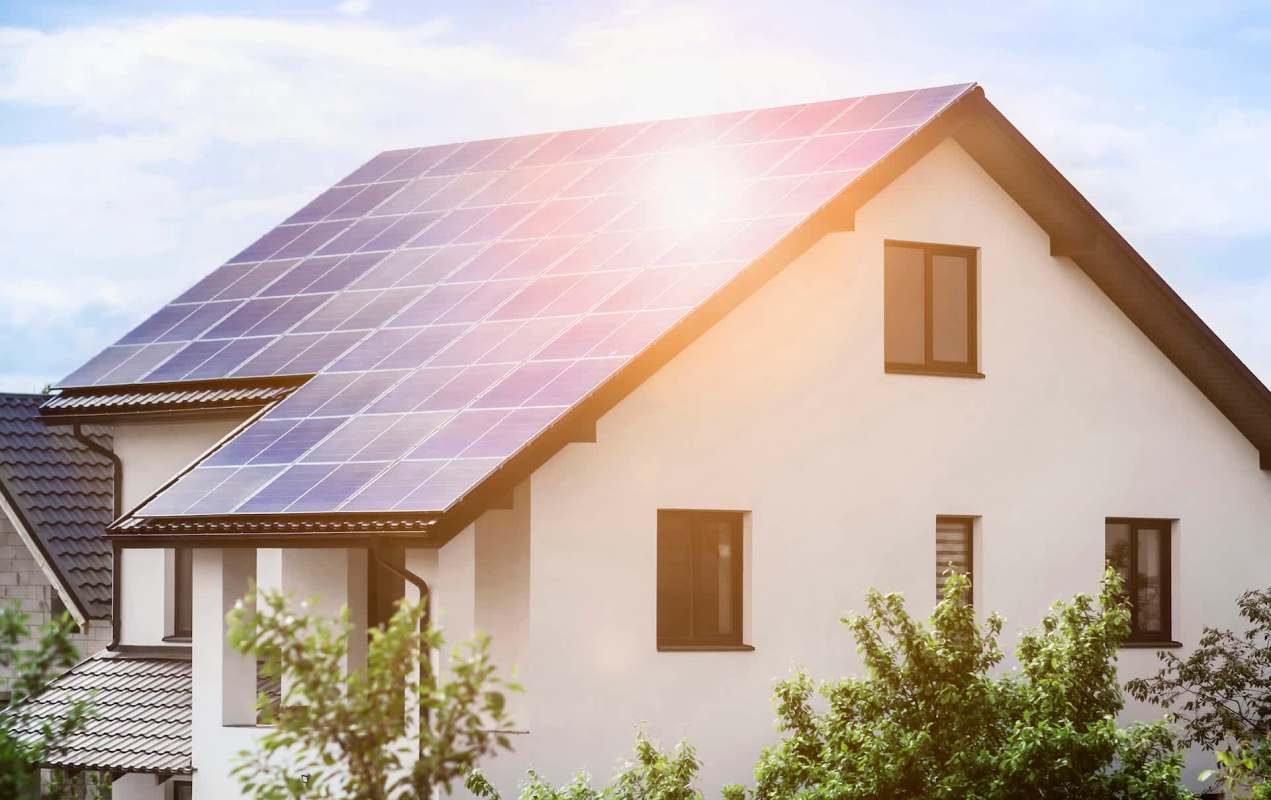 The sun shining brightly on a house with solar panels on the roof and surrounding greenery,