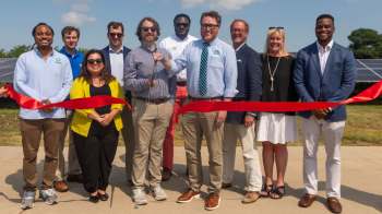 A group of people holding a red ribbon during a ribbon-cutting ceremony in front of solar panels.