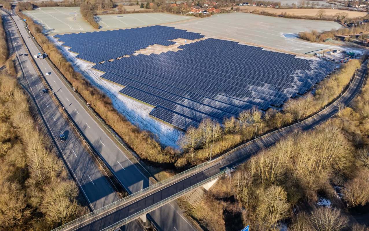 Aerial view of a large solar panel installation adjacent to a highway surrounded by trees.