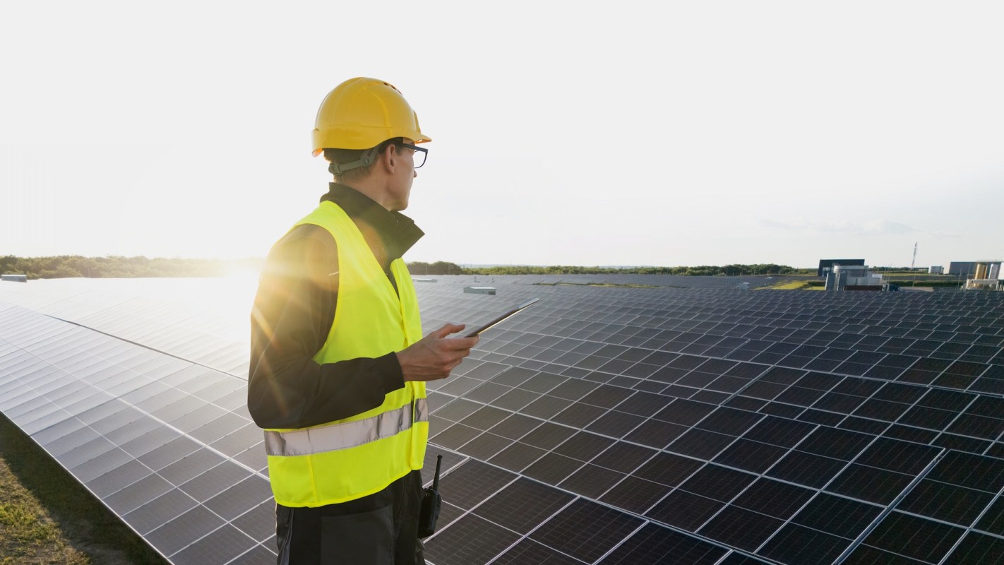 A worker in a safety vest and helmet stands near solar panels, holding a device.
