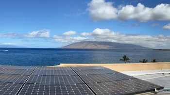 Solar panels on a roof overlooking a blue ocean and distant hills under a partly cloudy sky.