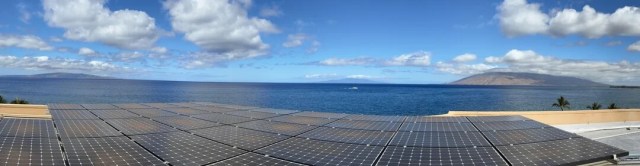 Solar panels on a roof overlooking a blue ocean and distant hills under a partly cloudy sky.