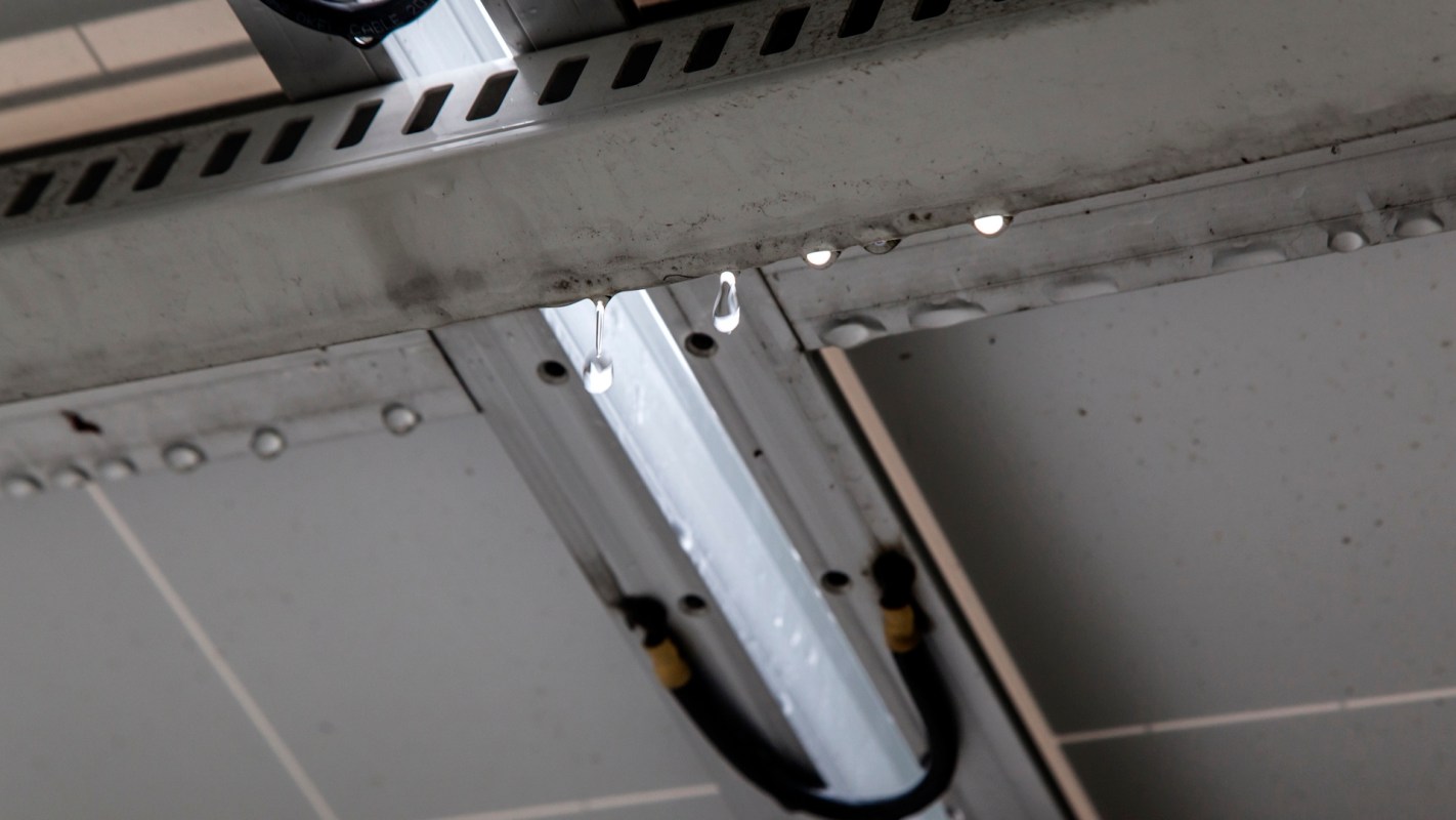 Water droplets hang from a metallic surface underneath a fluorescent light fixture.