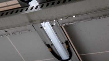 Water droplets hang from a metallic surface underneath a fluorescent light fixture.