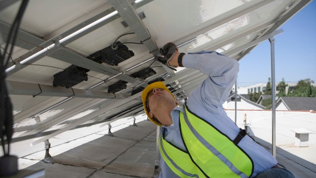 A worker in a safety vest repairs solar panels on a roof.