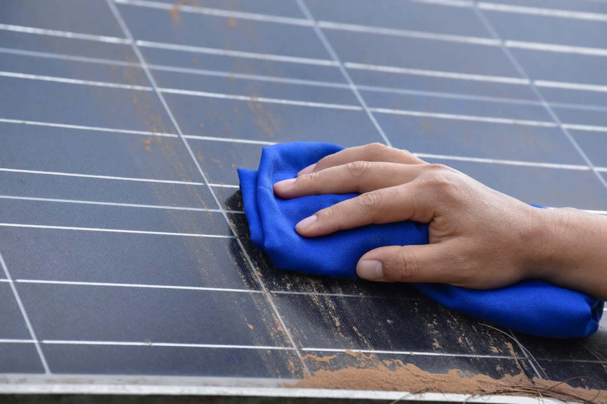 A hand holds a blue cloth while cleaning a dusty solar panel.