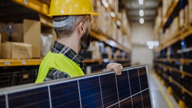 A person holds a solar panel in a warehouse with shelves stocked with boxes.