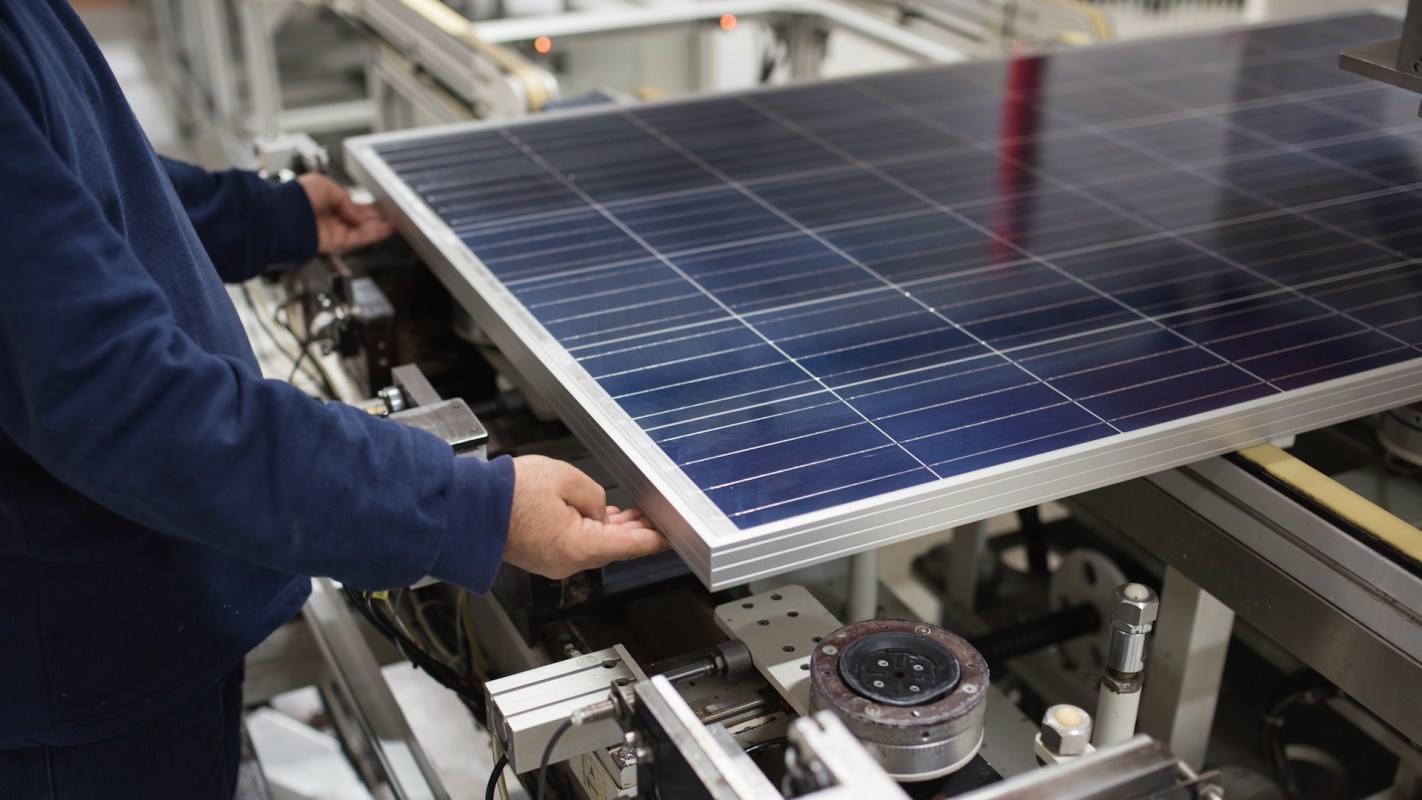A person handles a solar panel on a production line amid machinery and equipment.