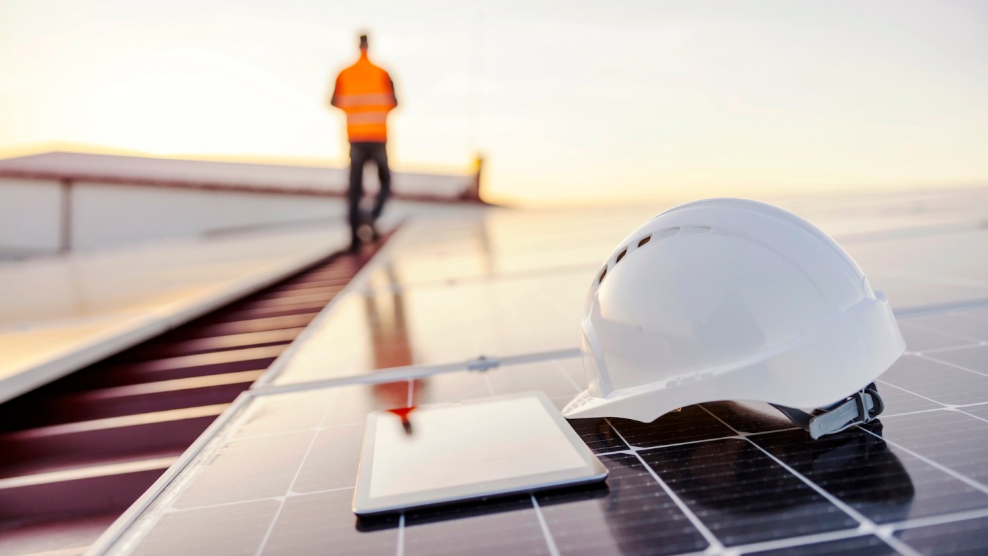 A hard hat and a tablet are placed on solar panels with a worker in the background.