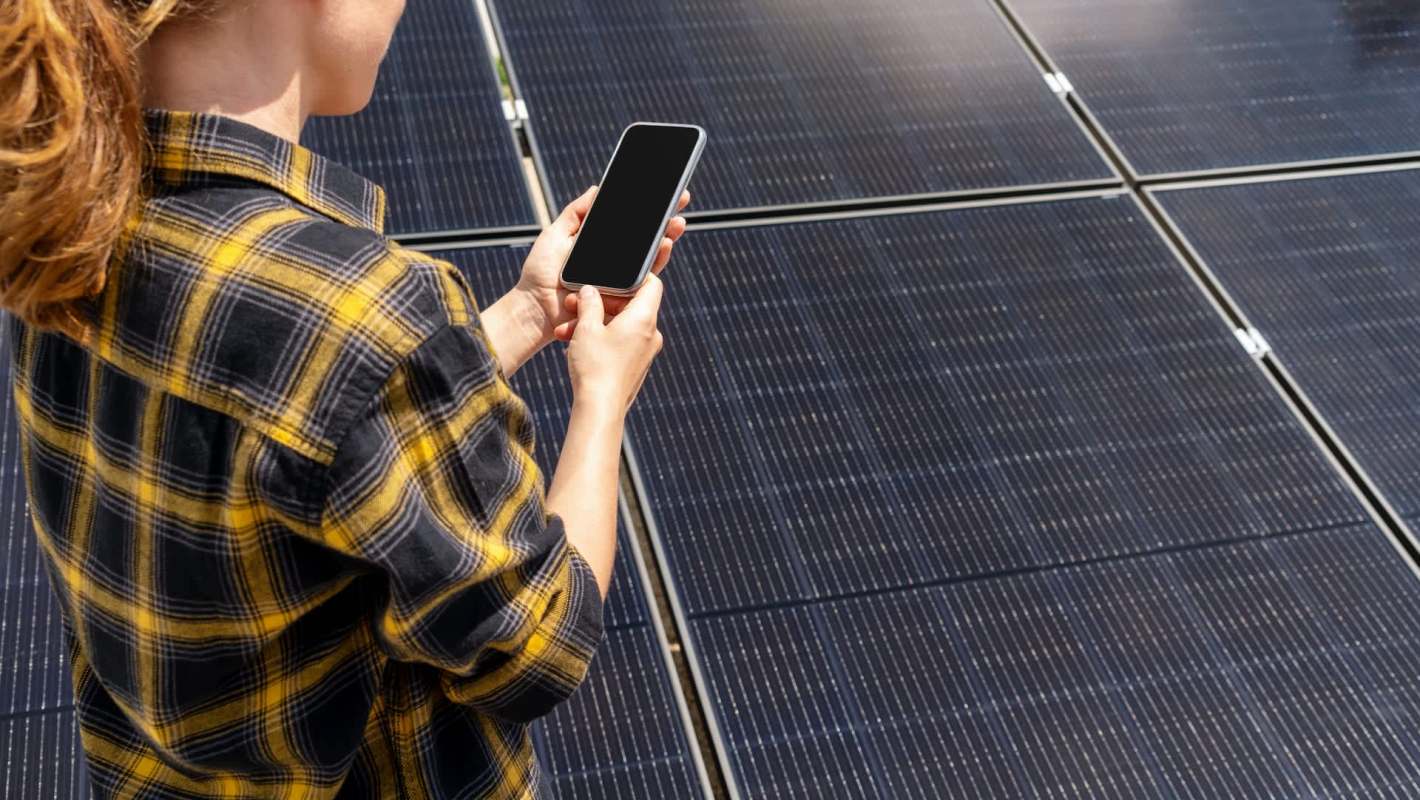 A person holds a smartphone while standing near solar panels.