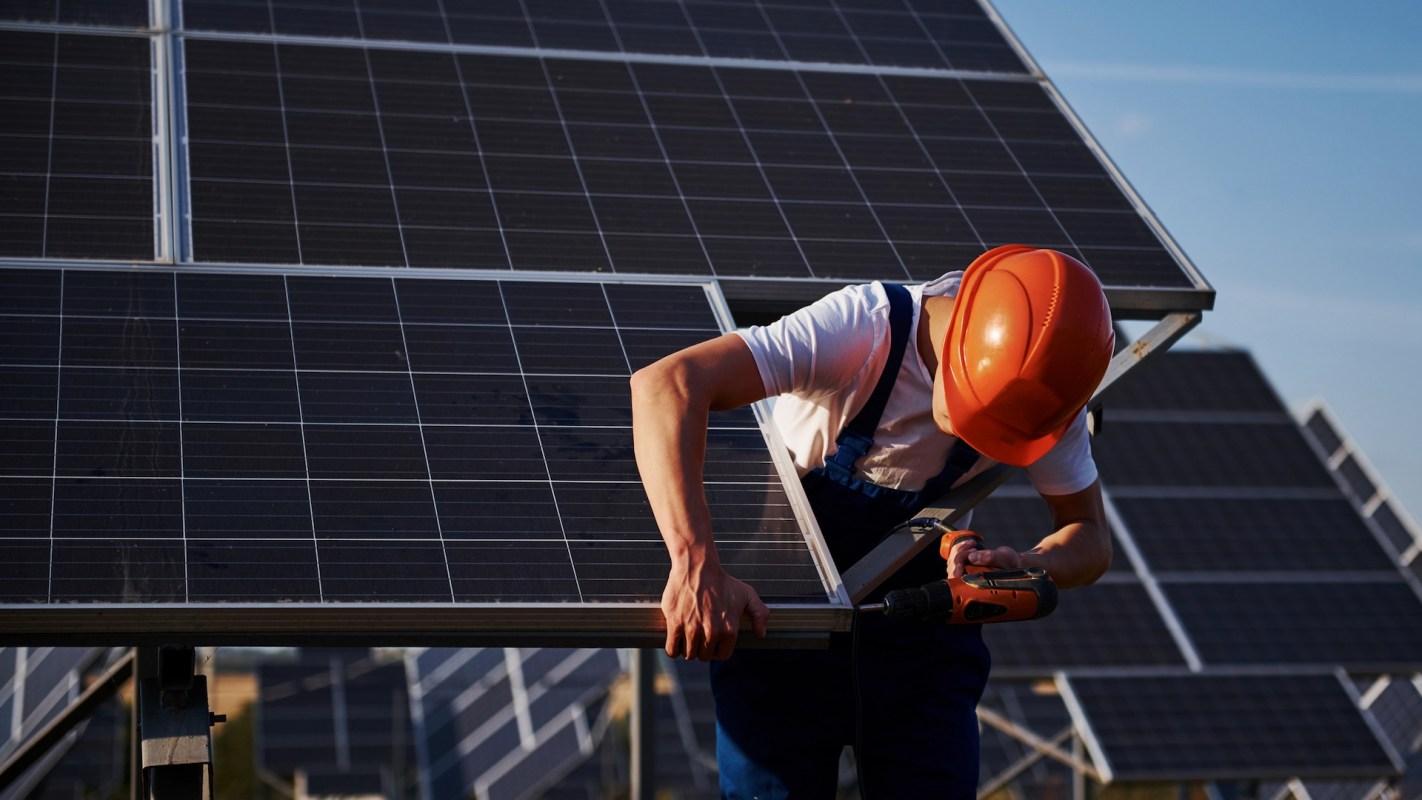A person in an orange helmet works on a solar panel with a power drill.