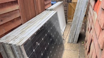 Solar panels leaning against a wooden fence in a narrow outdoor space.