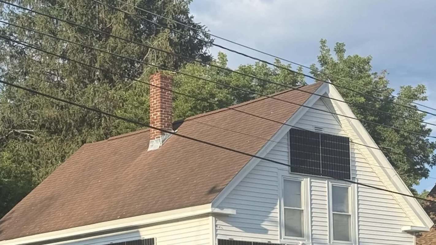 A house with a brown roof, chimney, and solar panels on the side, surrounded by trees and power lines.