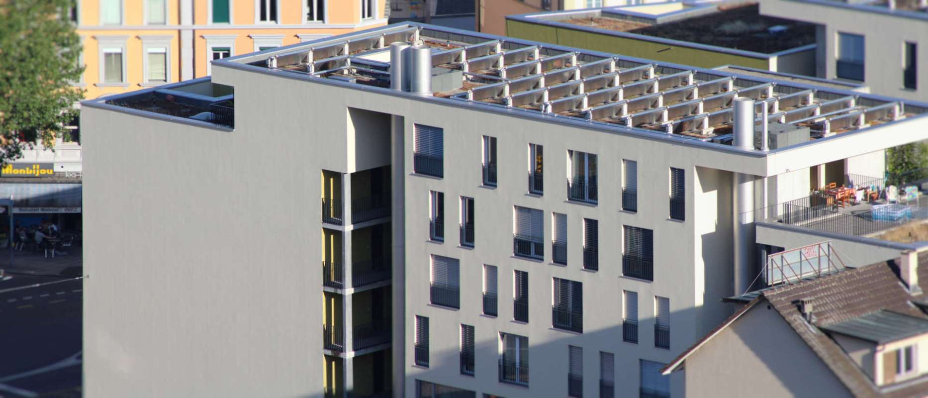 A modern building with balconies and a rooftop structure, located near a street with outdoor seating.