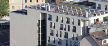 A modern building with balconies and a rooftop structure, located near a street with outdoor seating.