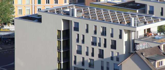 A modern building with balconies and a rooftop structure, located near a street with outdoor seating.