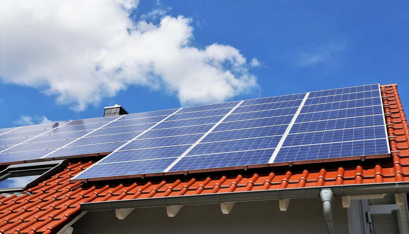 Solar panels installed on a red-tiled roof against a blue sky with clouds.