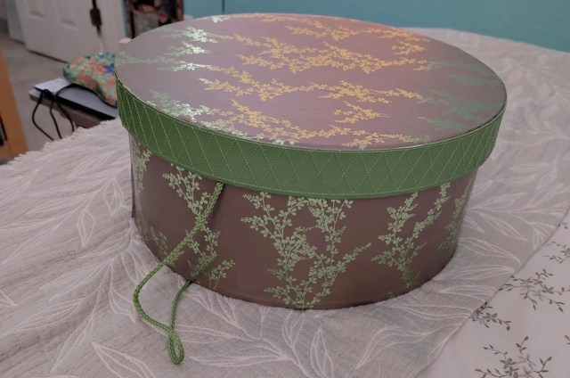 A round decorative box with a floral design on top and a green patterned band is placed on a textured surface.