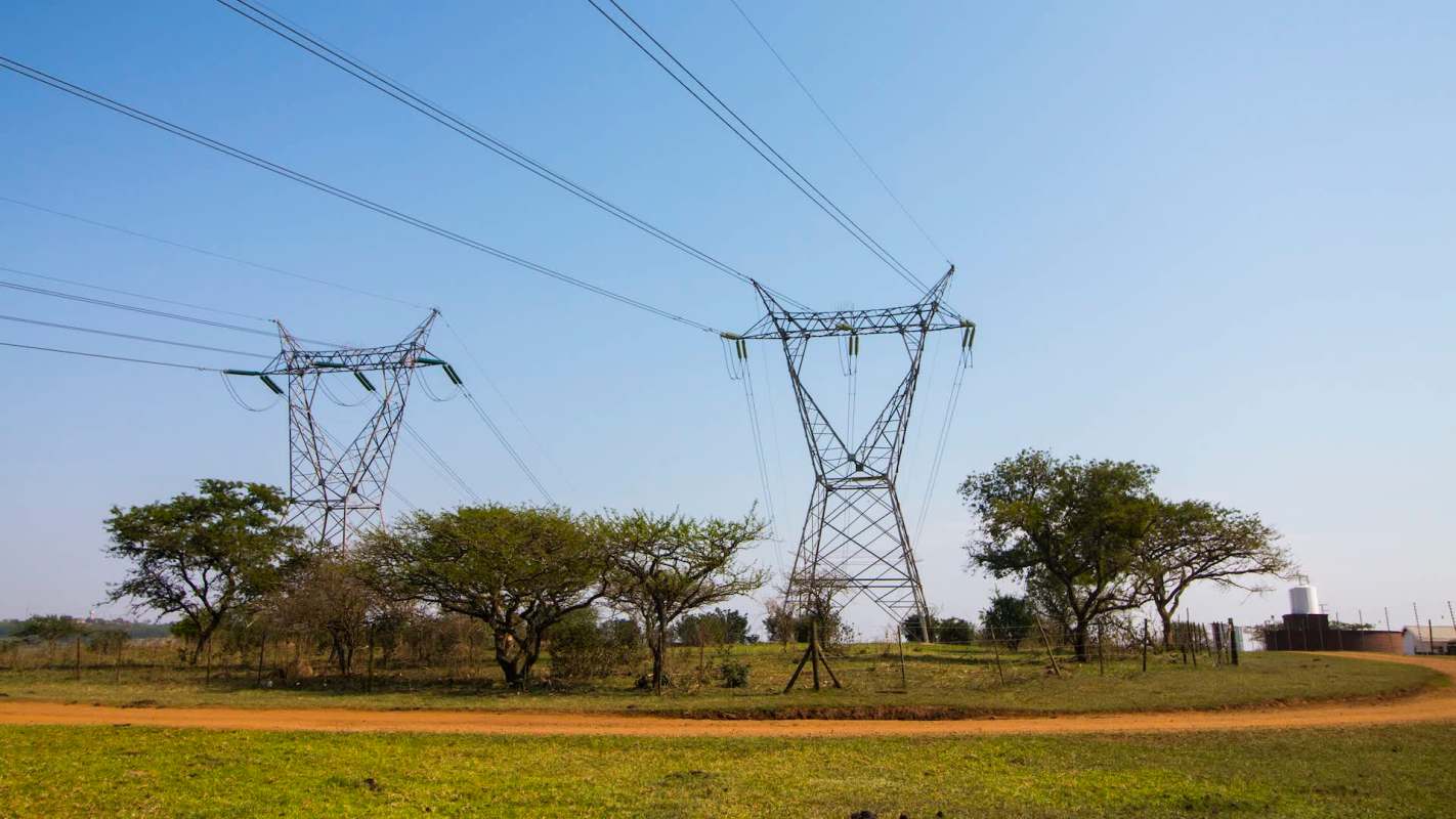 Two power transmission towers stand in an open field with trees and a dirt path.