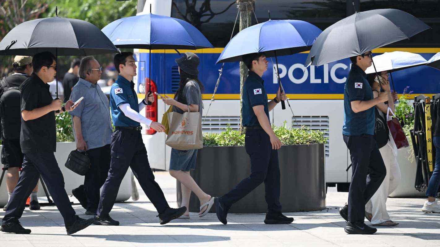 People walk on a street, some holding umbrellas, alongside a police van.