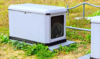 A gray outdoor standby generator with a fan, surrounded by grass and vegetation.
